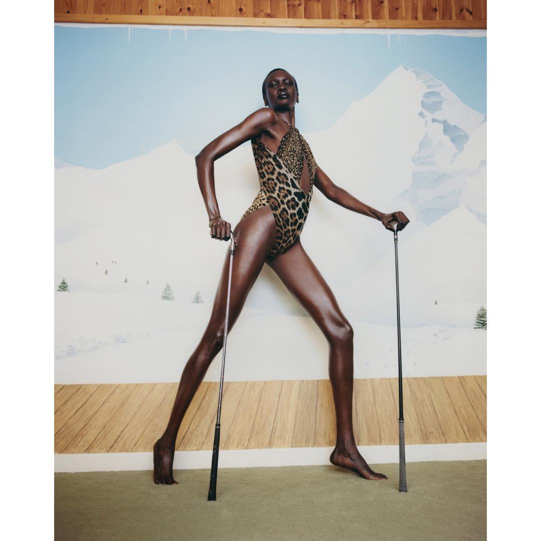 Fashionable woman in a leopard print bathing suit poses confidently with ski poles against a painted backdrop of snow-covered mountains.