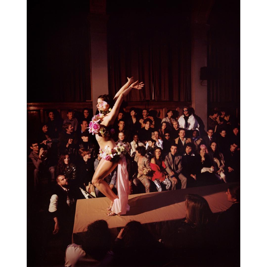 A fashion show featuring a performer adorned with floral embellishments on a stage, with an audience in the background, creating an editorial style image.