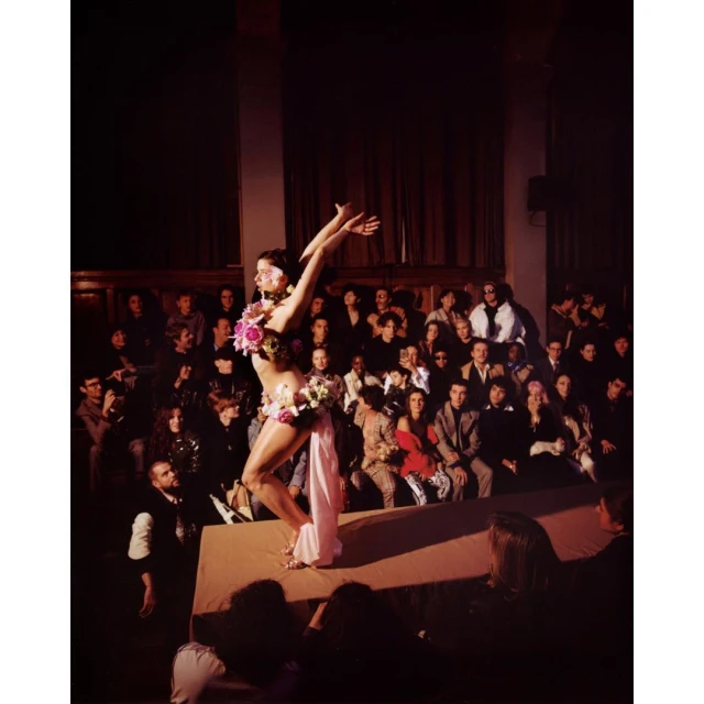 A fashion show featuring a performer adorned with floral embellishments on a stage, with an audience in the background, creating an editorial style image.