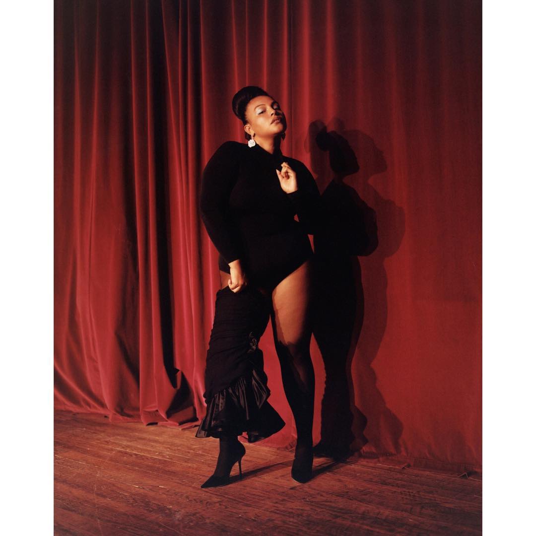 Portrait of a woman in a black leotard and stockings posing in front of a red curtain with a partial garment in her hand.