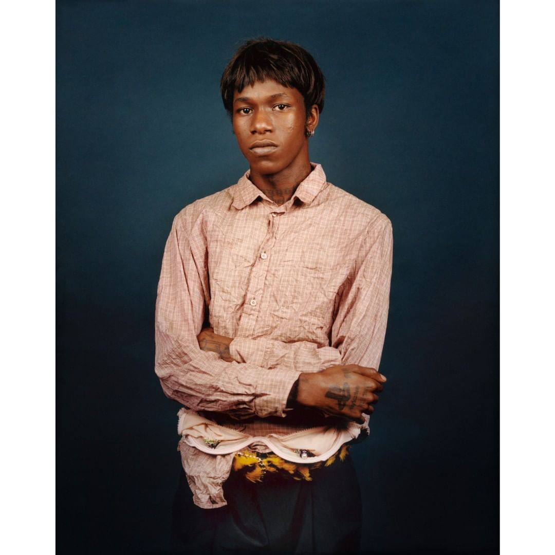 Portrait of a young adult with short dark hair, wearing a button-down shirt with arms crossed, standing against a dark blue background.