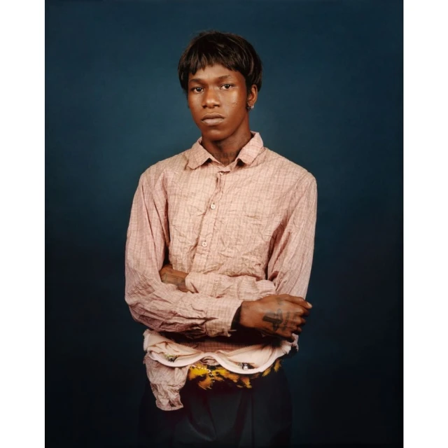Portrait of a young adult with short dark hair, wearing a button-down shirt with arms crossed, standing against a dark blue background.