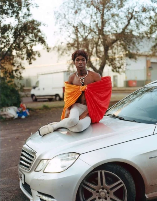 Person sitting on the hood of a silver Mercedes-Benz, wearing a red and orange draped garment, white tights, and white shoes in an urban setting.