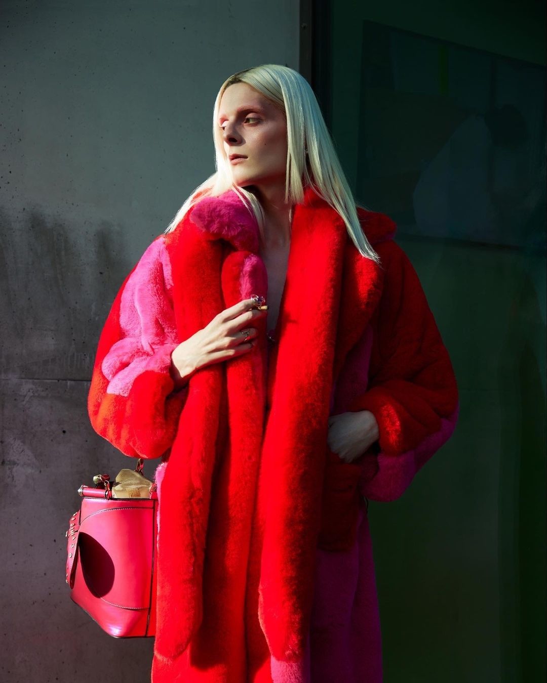 Fashionable adult in red faux fur coat and pink accessories stands against concrete wall in soft daylight.