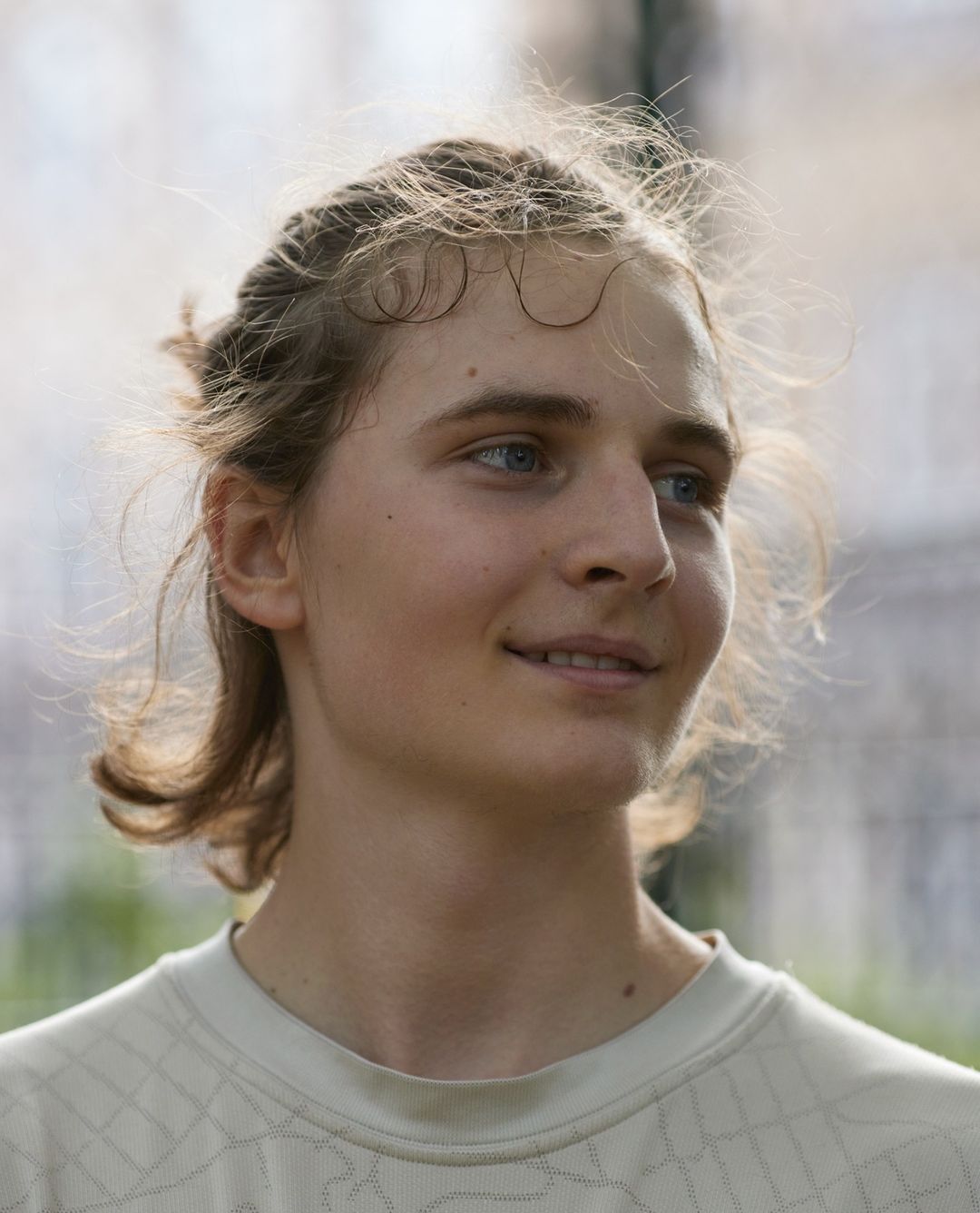 Close-up portrait of a young adult with light brown hair, fair skin, and blue eyes, wearing a light-colored patterned t-shirt outdoors.