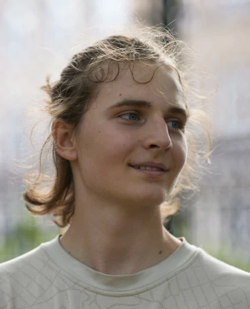Close-up portrait of a young person with light brown hair wearing a patterned off-white t-shirt against a soft outdoor background.