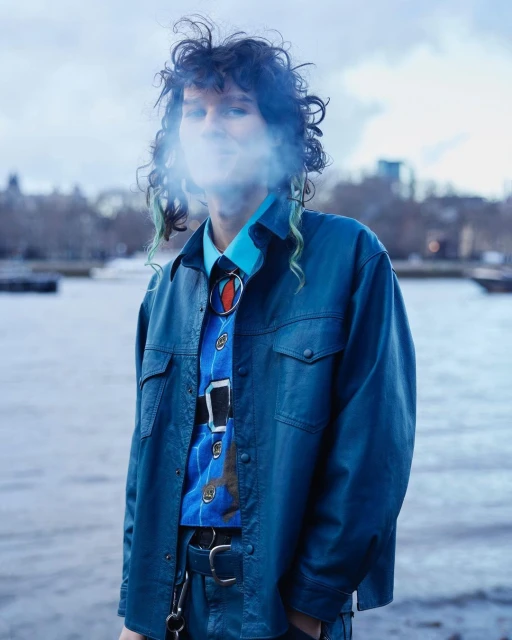 Stylish adult smokes in a blue jacket outdoors near a river, with buildings in the background, showcasing a unique and edgy fashion statement.