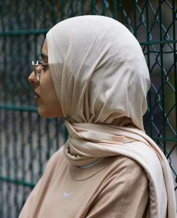 Profile portrait of a woman wearing a cream hijab and glasses against a green chain link fence