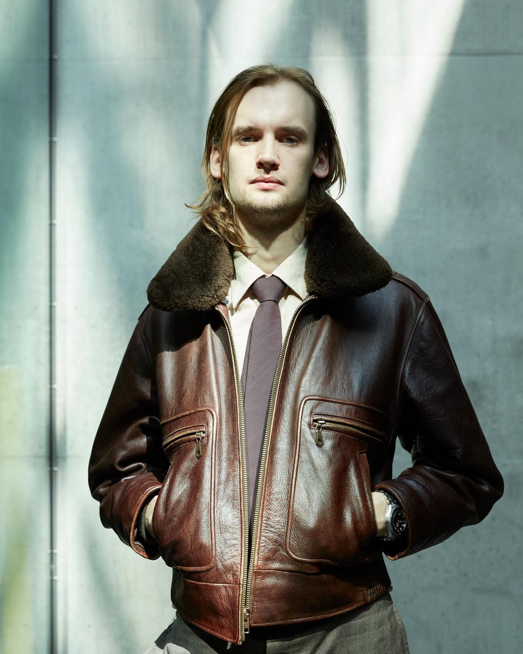 An adult male with long brown hair poses outdoors wearing a leather jacket with a fur collar, a shirt, and a tie, with hands in pockets.