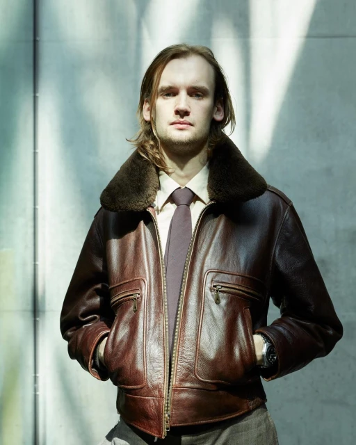 An adult male with long brown hair poses outdoors wearing a leather jacket with a fur collar, a shirt, and a tie, with hands in pockets.