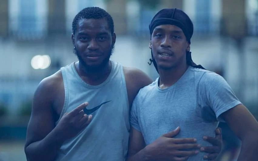 Two African-descent men pose outdoors in casual wear against an urban backdrop with neutral tones and soft, diffused natural lighting.