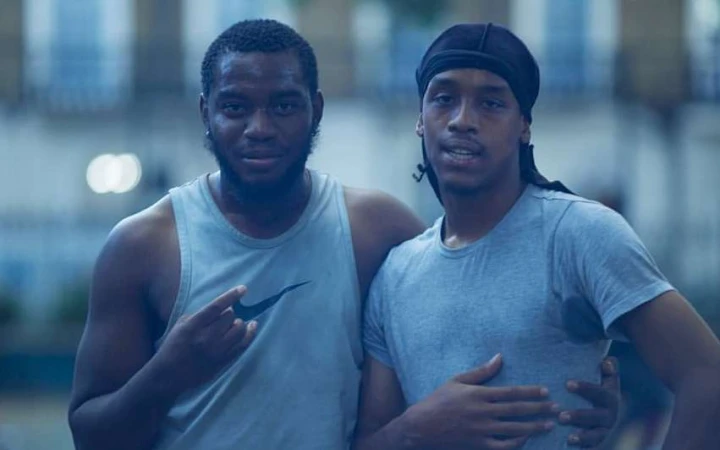Two African-descent men pose outdoors in casual wear against an urban backdrop with neutral tones and soft, diffused natural lighting.