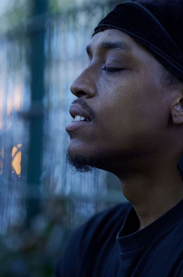 Profile of African American man with eyes closed wearing a black headscarf, T-shirt against a blurred urban backdrop. Focus on relaxation.