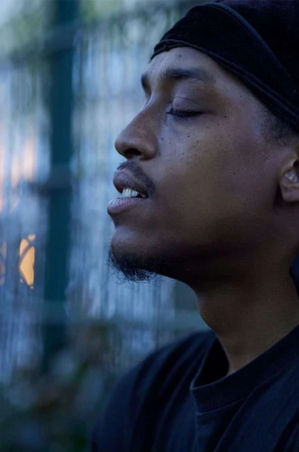 Profile of African American man with eyes closed wearing a black headscarf, T-shirt against a blurred urban backdrop. Focus on relaxation.