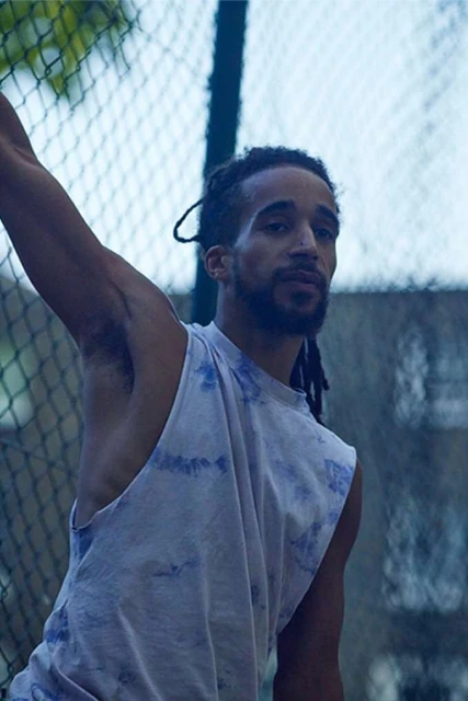 Athletic man with dreadlocks in tank top stands near chain-link fence outdoors, looking towards the camera.