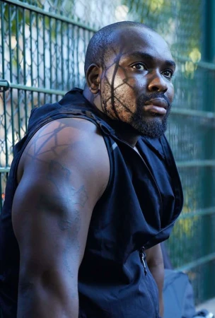 Portrait of a Black man with patterned shadow across his face wearing a black sleeveless top against a green wire fence by Olivia Beasley