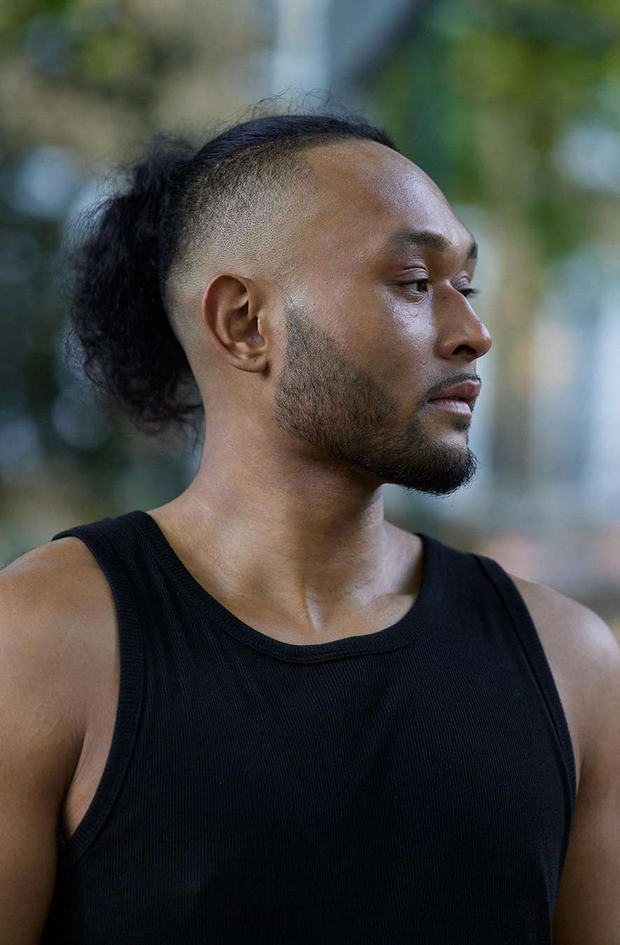 Portrait of a dark-skinned adult male with shaved sides and hair tied back, wearing a black tank top against a blurred background.
