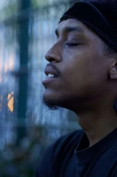 Profile portrait of a Black man with eyes closed wearing a black durag and dark t-shirt outdoors.