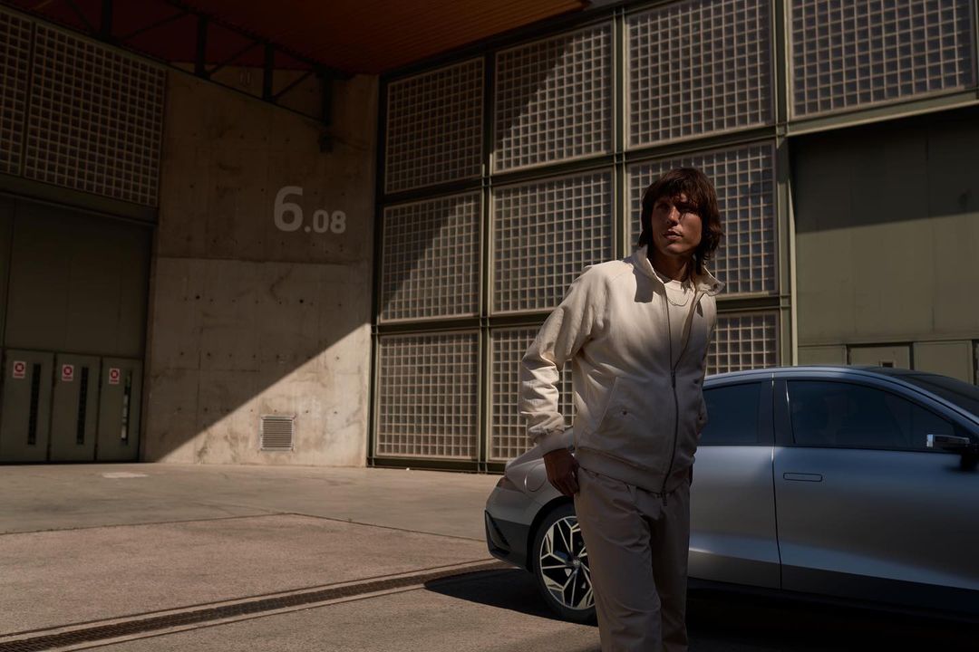 Adult male in a beige track suit standing in front of a silver car and building with the number 6.08, in an urban outdoor setting.