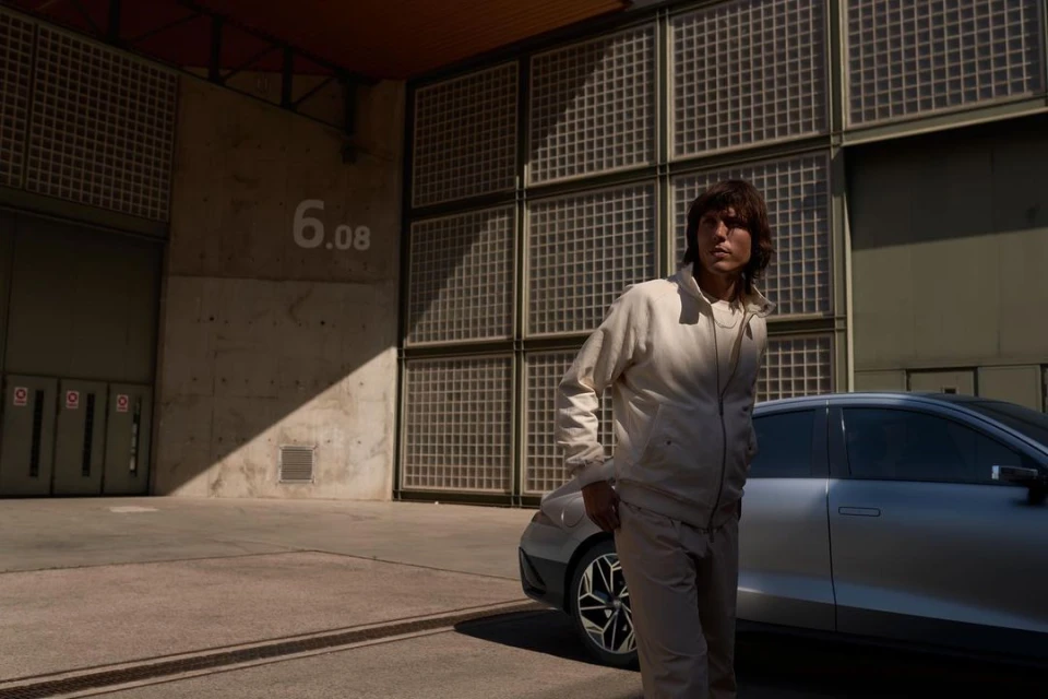 Adult male in a beige track suit standing in front of a silver car and building with the number 6.08, in an urban outdoor setting.
