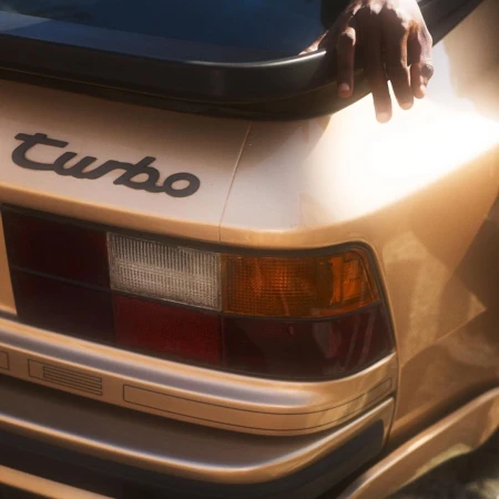 Rear view of a gold car with a spoiler and the word Turbo in cursive on the trunk, showing the tail light and a hand on the frame.