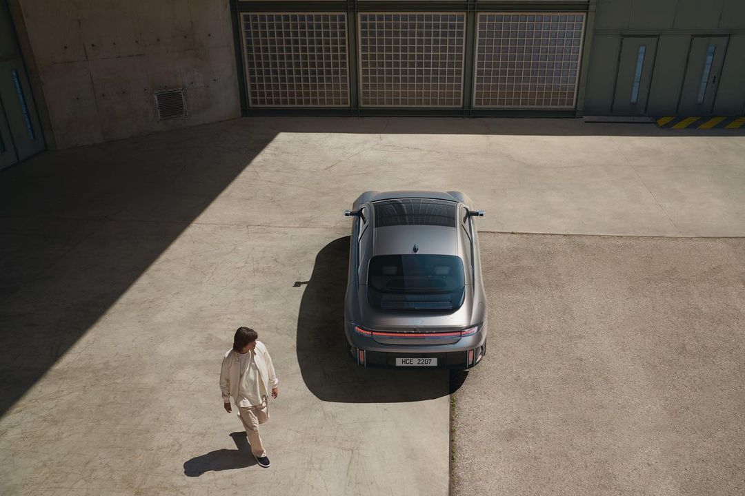 Aerial shot of a grey electric sedan parked on concrete with an adult in beige walking nearby, bathed in bright, natural light.