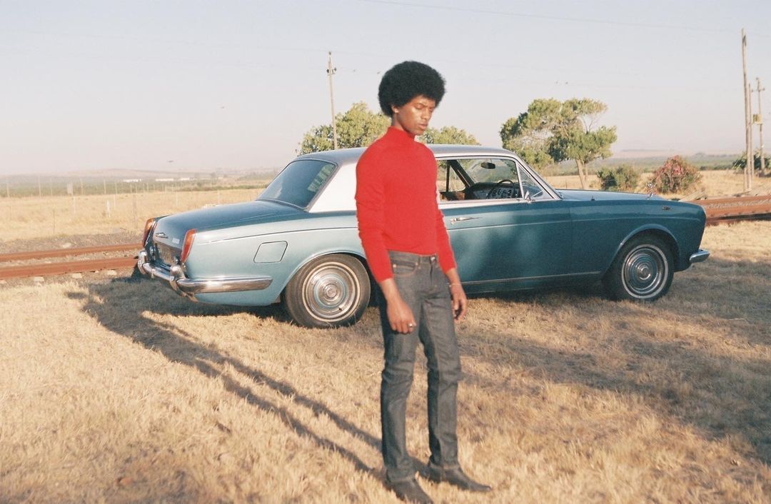 A person in a red turtleneck stands in a field next to a light blue vintage car with a white roof, capturing a moment in natural light.