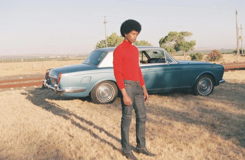 A person in a red turtleneck stands in a field next to a light blue vintage car with a white roof, capturing a moment in natural light.