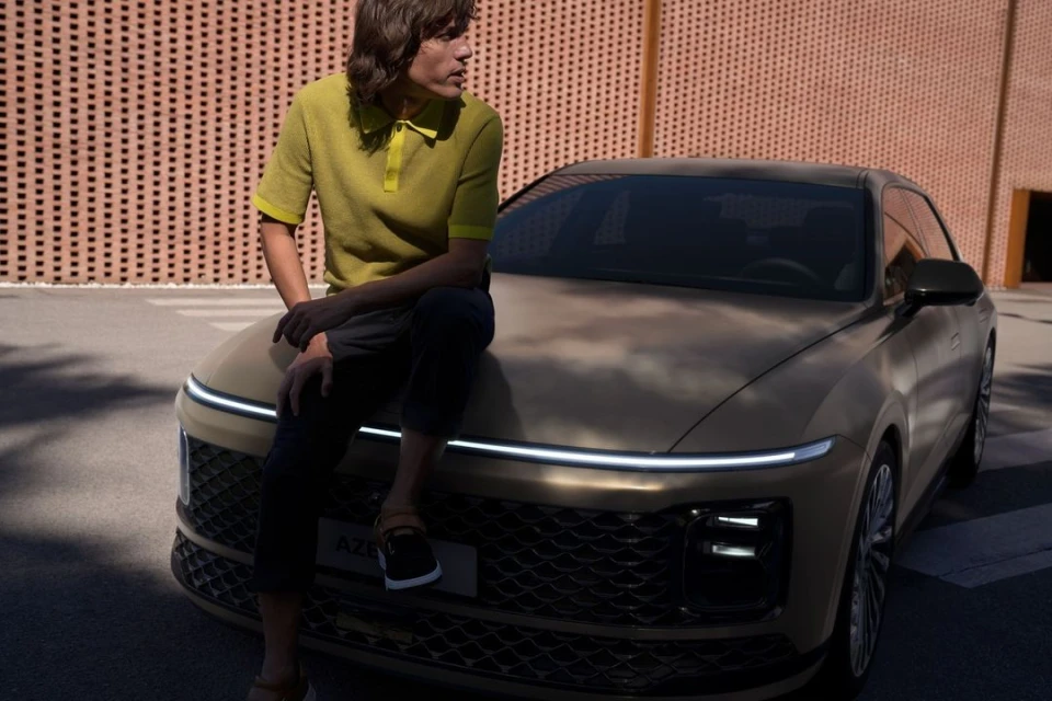An adult male model poses sitting on the hood of a modern, tan sedan car in front of a brick building, automotive fashion shoot.