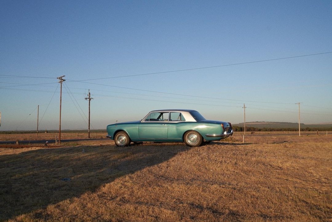 Side view of a teal vintage Rolls Royce parked on dry grassy field against a clear blue sky on a bright, sunny day in the countryside.