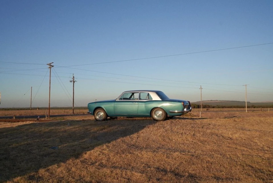 Side view of a teal vintage Rolls Royce parked on dry grassy field against a clear blue sky on a bright, sunny day in the countryside.
