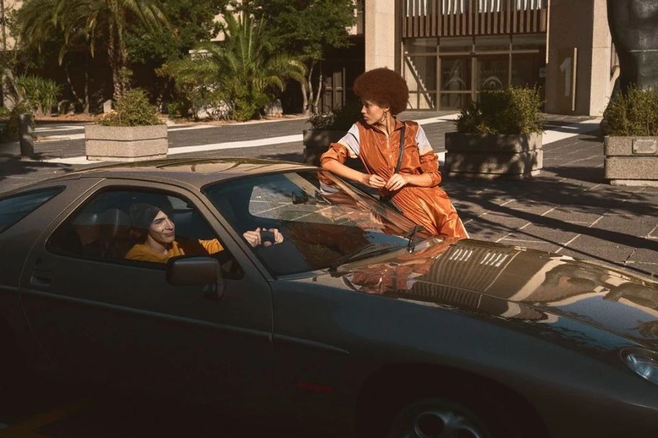 A vintage grey Porsche parked outdoors on a sunny day, featuring an adult male driver and an adult female passenger leaning on the car.