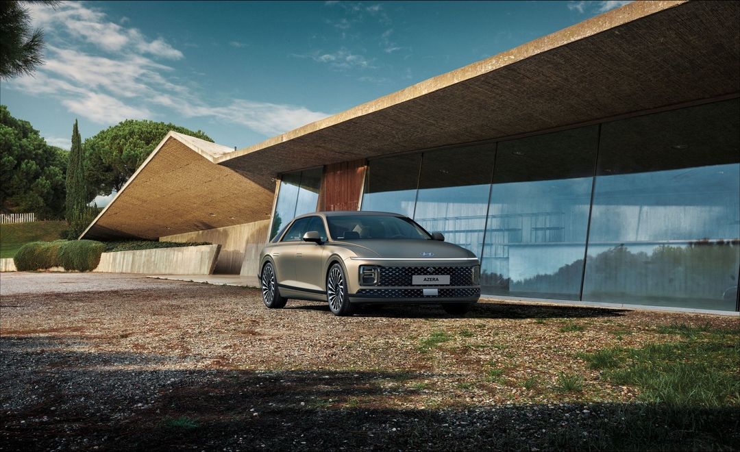 Champagne-colored Hyundai Azera parked in front of a modern building with large windows, gravel ground, trees and blue sky with clouds.