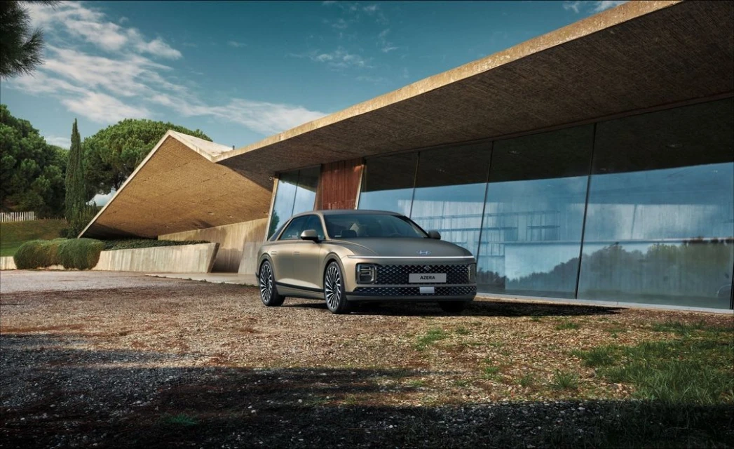 Champagne-colored Hyundai Azera parked in front of a modern building with large windows, gravel ground, trees and blue sky with clouds.