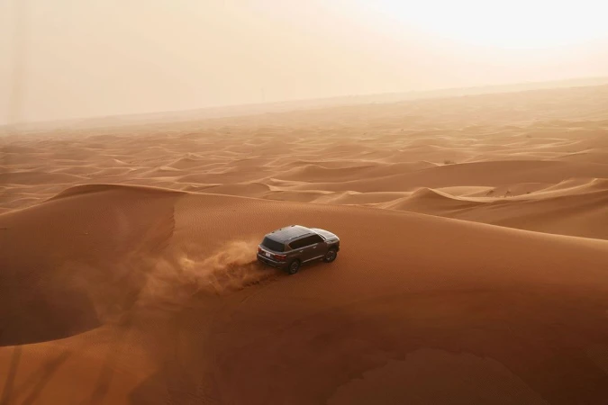 SUV vehicle driving on sand dunes in desert environment with warm sunny light and sandy color palette, transport, landscape photography.