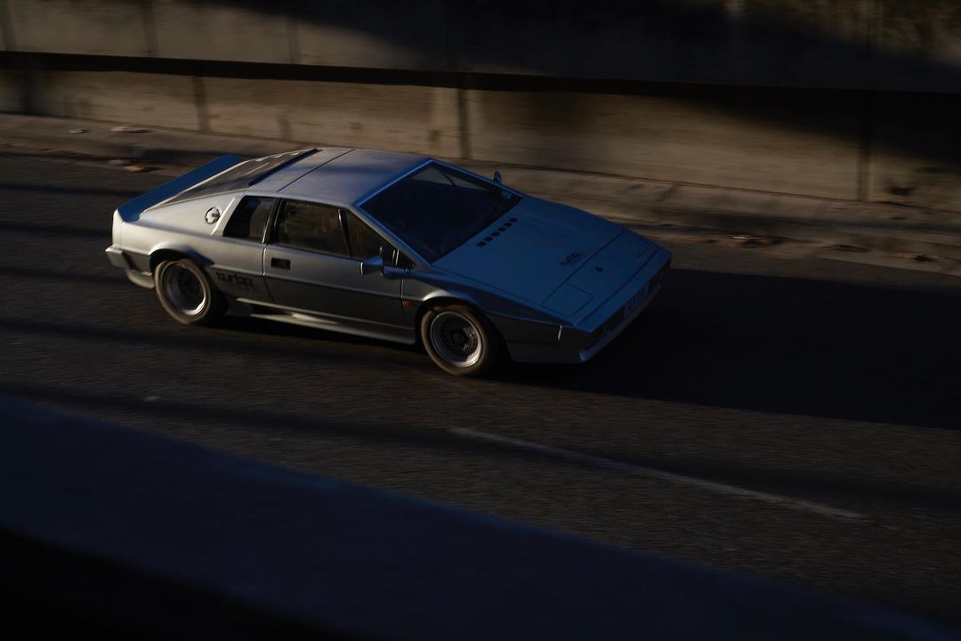 Silver Lotus Esprit driving on a sunny road with a concrete barrier in the background, highlighting its sleek design and speed.
