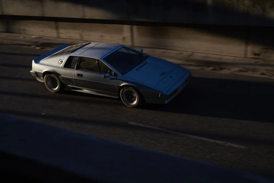 Silver Lotus Esprit driving on a sunny road with a concrete barrier in the background, highlighting its sleek design and speed.