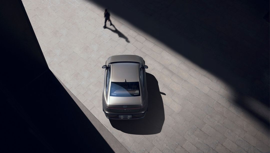 Aerial shot of a silver sedan parked on a textured grey surface, with a single figure casting a shadow in the background.