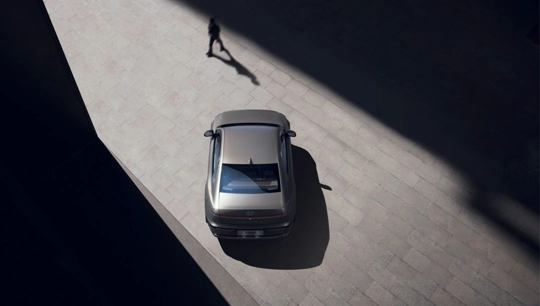 Aerial shot of a silver sedan parked on a textured grey surface, with a single figure casting a shadow in the background.