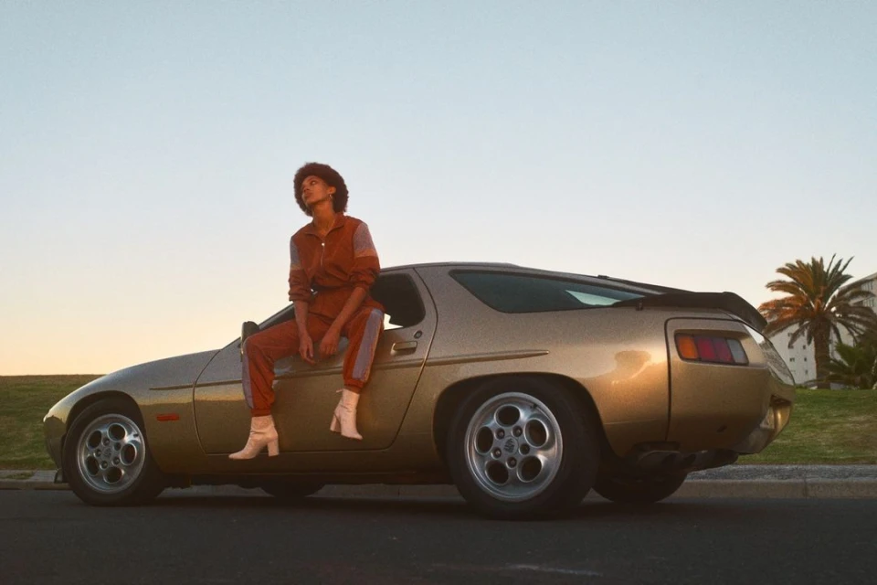 A woman sits on the gold hood of a vintage Porsche 928 under the golden sunset sky, wearing a brown jumpsuit and white boots.