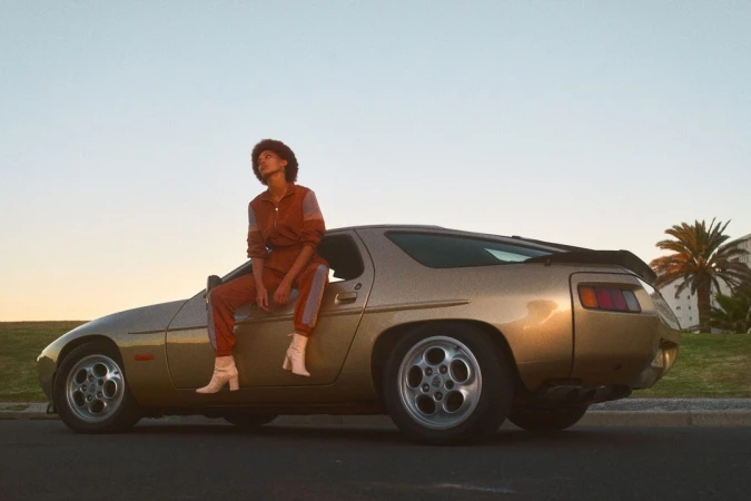 A woman sits on the gold hood of a vintage Porsche 928 under the golden sunset sky, wearing a brown jumpsuit and white boots.