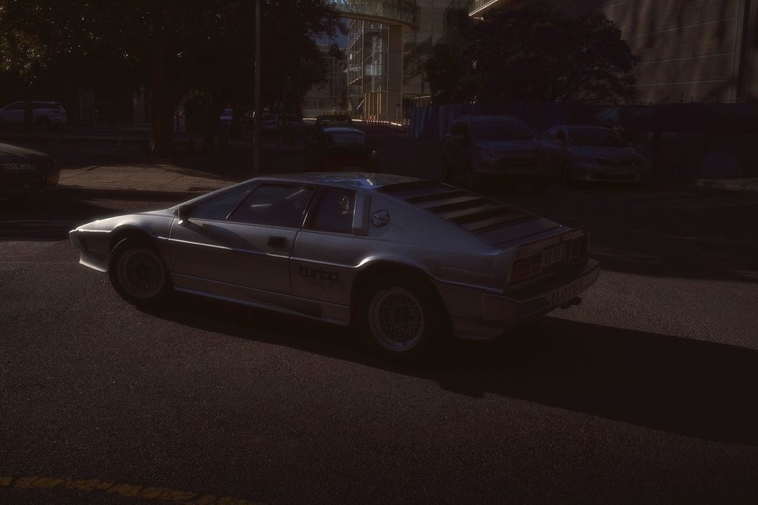 Silver Lotus Esprit Turbo parked on a city street with a dark background and a contemporary building during daylight hours.