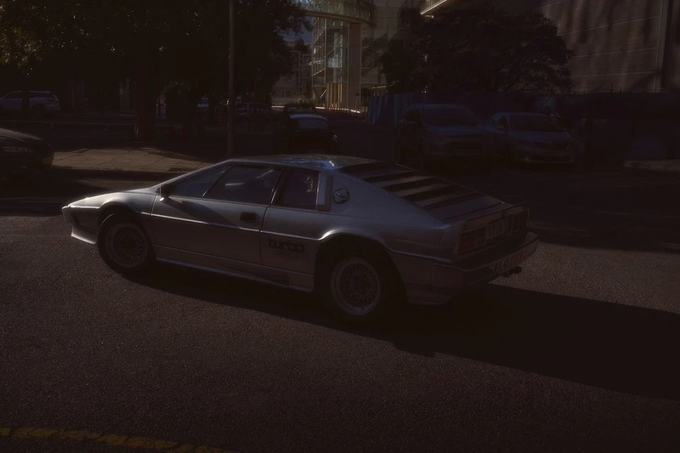 Silver Lotus Esprit Turbo parked on a city street with a dark background and a contemporary building during daylight hours.