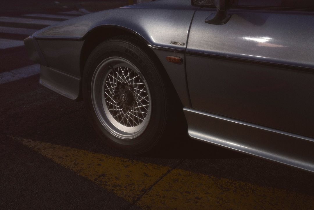 Silver sports car with multi-spoke wheel and low-profile tire parked on a striped surface with a hard-shadow yellow stripe.