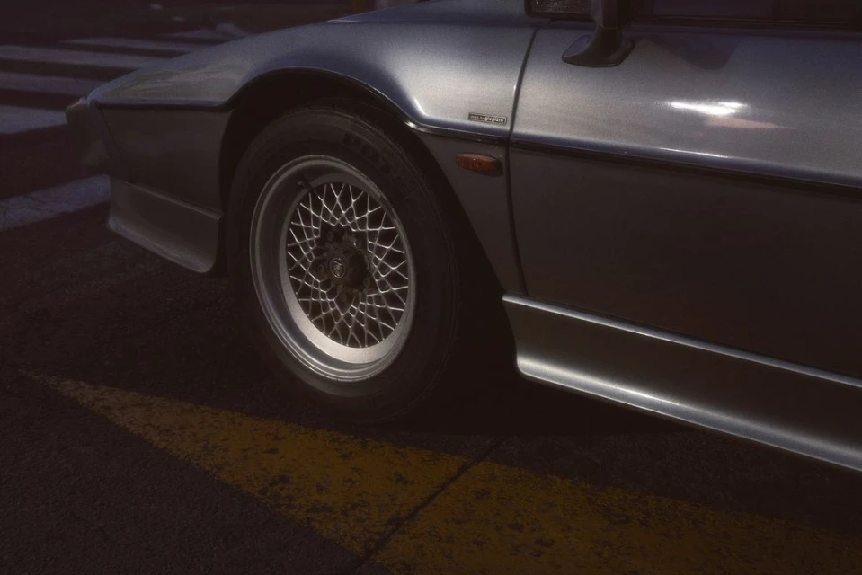Silver sports car with multi-spoke wheel and low-profile tire parked on a striped surface with a hard-shadow yellow stripe.