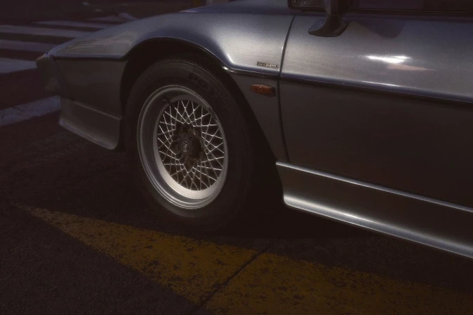 Silver sports car with multi-spoke wheel and low-profile tire parked on a striped surface with a hard-shadow yellow stripe.