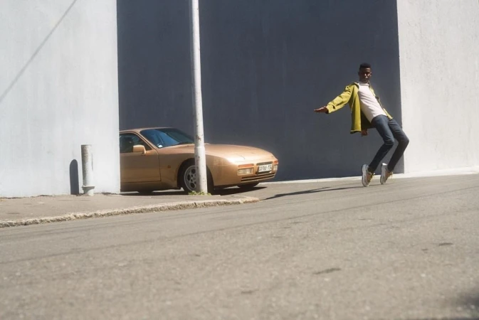 Man in yellow jacket leaning back on a sloping street next to a parked gold sports car