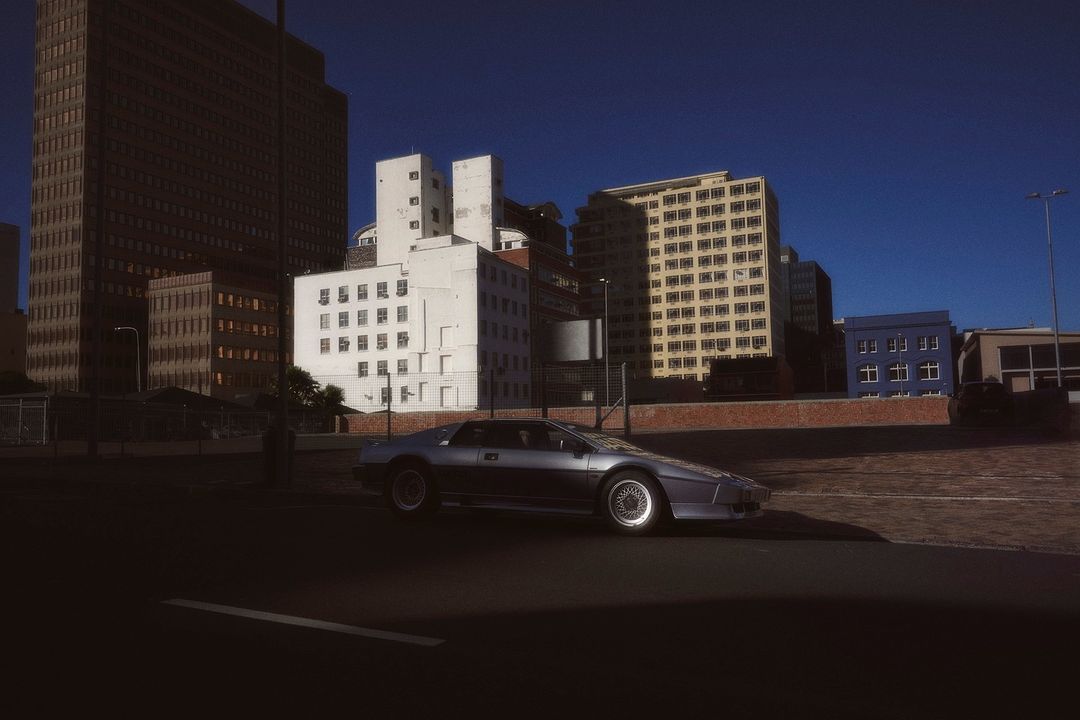 Silver coupe parked in an urban setting with tall buildings, under a clear blue sky on a sunny day casting shadows.