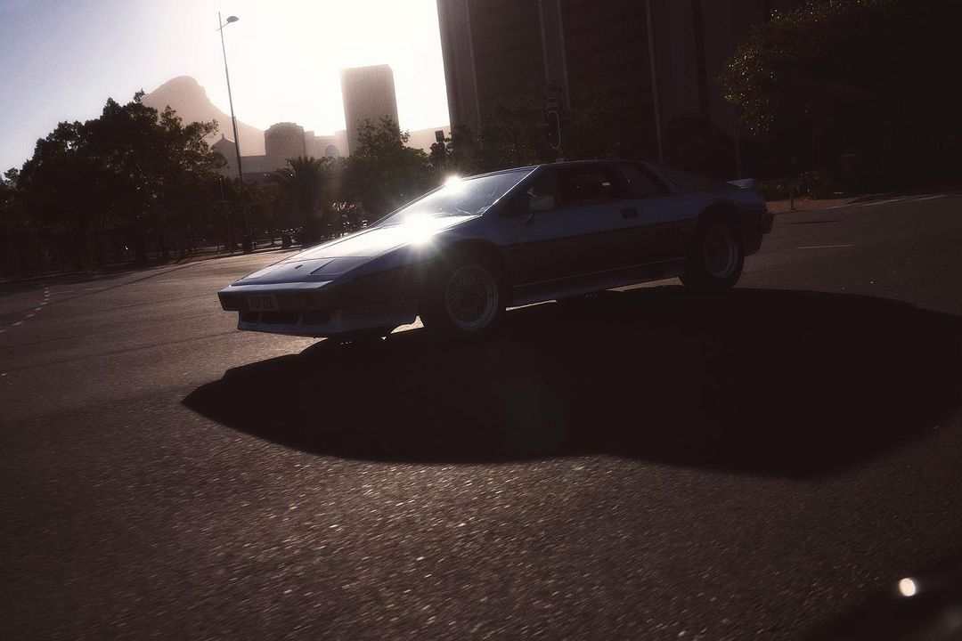 Dark blue sportscar is parked on an asphalt road on a bright day. Trees, city buildings, and a mountain are in the background.