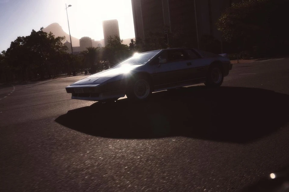 Dark blue sportscar is parked on an asphalt road on a bright day. Trees, city buildings, and a mountain are in the background.